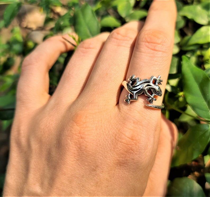 May include: A silver-colored ring in the shape of a gecko, with detailed scales and limbs. The ring wraps around the finger, creating an adjustable fit. The gecko ring is a unique piece of jewelry.