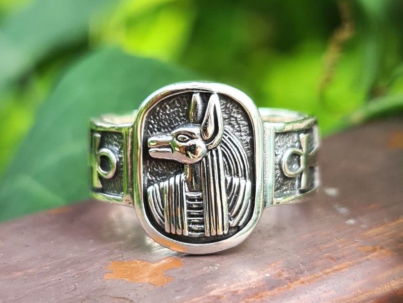 May include: Silver ring with an Anubis head design. The ring is set in a rectangular frame with an ankh symbol on each side.
