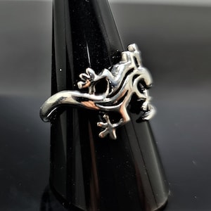 May include: A silver-colored ring featuring a stylized lizard design. The ring has a detailed, three-dimensional lizard motif with intricate patterns and textures. The lizard is posed in a dynamic, crawling position, with its legs and tail curved around the ring's band.