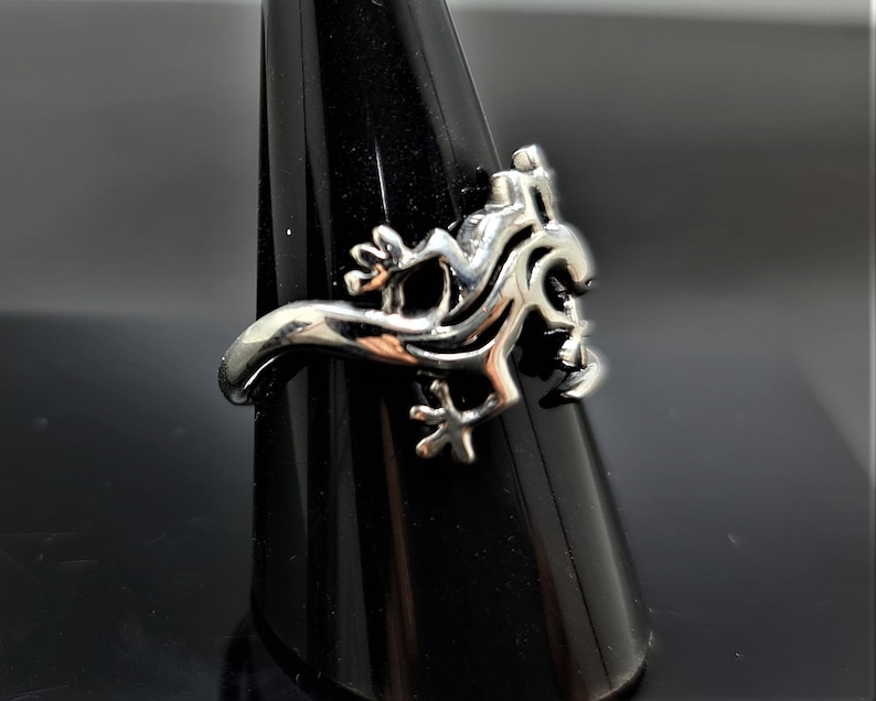 May include: A silver-colored ring featuring a stylized lizard design. The ring has a detailed, three-dimensional lizard motif with intricate patterns and textures. The lizard is posed in a dynamic, crawling position, with its legs and tail curved around the ring's band.