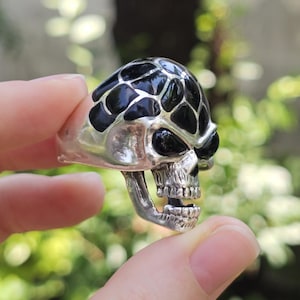 May include: A silver skull ring with a black, patterned top and black eye sockets. The skull's mouth is open, revealing teeth. The ring is held between fingers against a blurred green background.