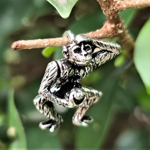 May include: A silver charm of a monkey sitting on a branch, with its arms raised above its head.