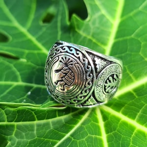 May include: A silver ring with intricate designs, featuring a circular pattern with a raised center and a smaller circular pattern with a tree-like design on the side.