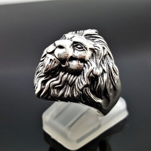 May include: A silver ring with a detailed lion head design. The lion's head is facing to the side and has a mane of hair.