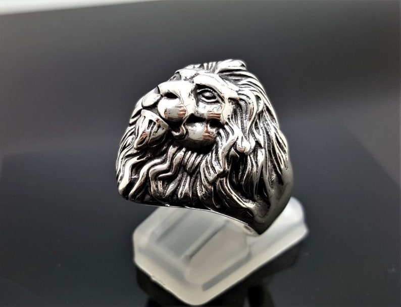 May include: A silver ring with a detailed lion head design. The lion's head is facing to the side and has a mane of hair.