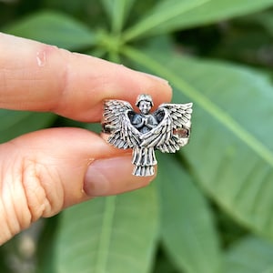 May include: Silver ring featuring a detailed angel design with outstretched wings. The angel is depicted with a serene expression and hands clasped in prayer.