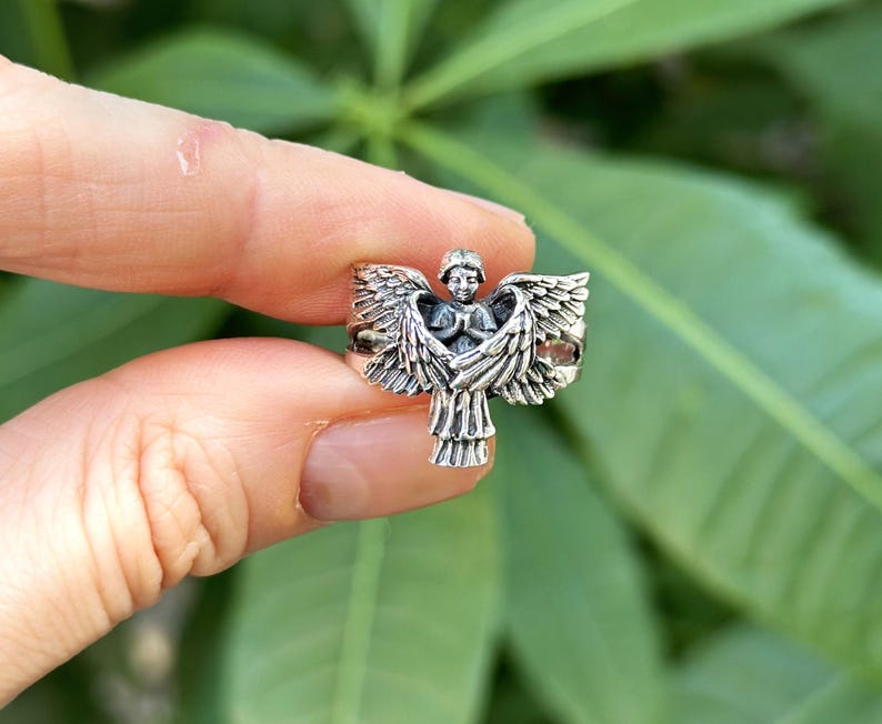 May include: Silver ring featuring a detailed angel design with outstretched wings. The angel is depicted with a serene expression and hands clasped in prayer.