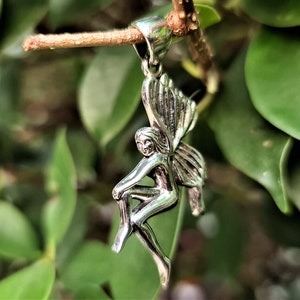 May include: A silver charm pendant featuring a detailed fairy with wings, sitting with one leg bent. The charm is hanging from a branch.