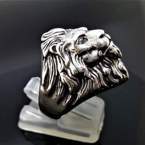 May include: A silver ring with a detailed lion head design. The lion's head is facing to the right and has a textured mane.
