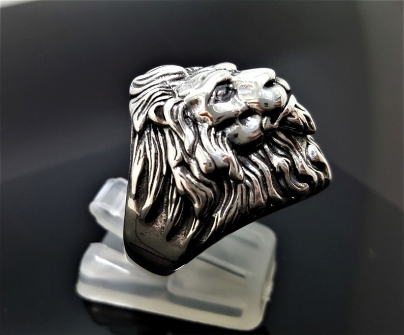 May include: A silver ring with a detailed lion head design. The lion's head is facing to the right and has a textured mane.