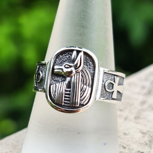 May include: A silver ring with an ankh symbol on one side and an image of the Egyptian goddess Bastet on the other side.