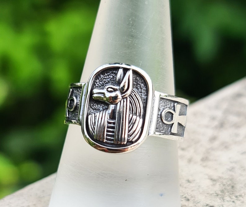 May include: A silver ring with an ankh symbol on one side and an image of the Egyptian goddess Bastet on the other side.