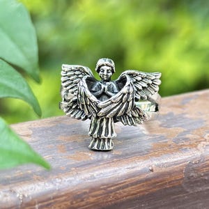 May include: Silver ring featuring a detailed angel design with outstretched wings. The angel is depicted with hands clasped in prayer.