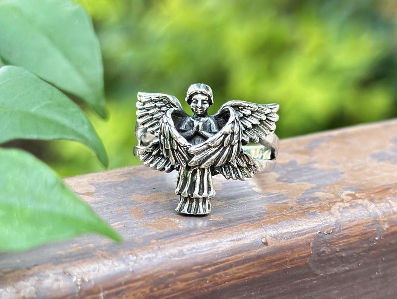 May include: Silver ring featuring a detailed angel design with outstretched wings. The angel is depicted with hands clasped in prayer.