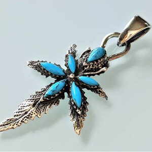 Pode incluir: Um pingente de prata com um design de flor turquesa. O pingente tem a forma de uma cruz com detalhes intrincados e um aro para prender uma corrente.