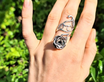 May include: A silver ring with a rose design. The ring is worn on the middle finger of a hand.