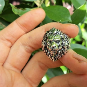 May include: A silver ring with a detailed lion head design. The lion's mane is intricately sculpted and the ring is worn on a finger.