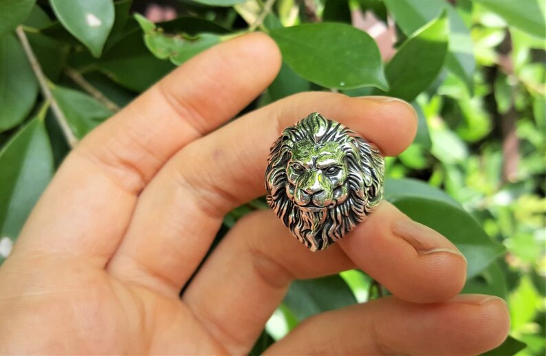 May include: A silver ring with a detailed lion head design. The lion's mane is intricately sculpted and the ring is worn on a finger.