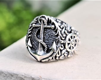 May include: A silver ring with an anchor and a ship's wheel design. The anchor is detailed with rope and the ring has a textured surface.