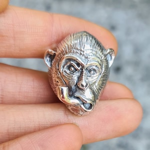 Puede incluir: Un anillo de plata detallado con forma de cabeza de mono, con una pipa. El anillo tiene intrincados rasgos faciales y una superficie texturizada. La expresión del mono es severa. Una pieza de joyería llamativa.