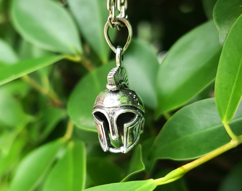 May include: Silver pendant necklace featuring a detailed depiction of a Roman soldier's helmet. The helmet is silver with a raised crest and intricate details. The necklace is hanging against a backdrop of green leaves.