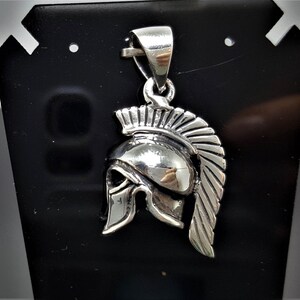 May include: Silver pendant featuring a detailed depiction of a Spartan helmet with a plume. The helmet is crafted in a realistic style with intricate details.