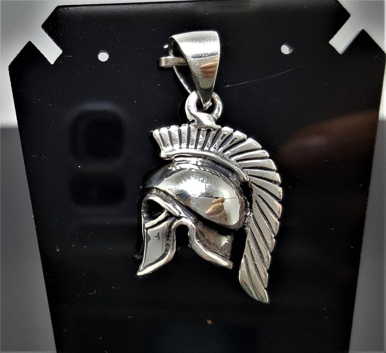 May include: Silver pendant featuring a detailed depiction of a Spartan helmet with a plume. The helmet is crafted in a realistic style with intricate details.