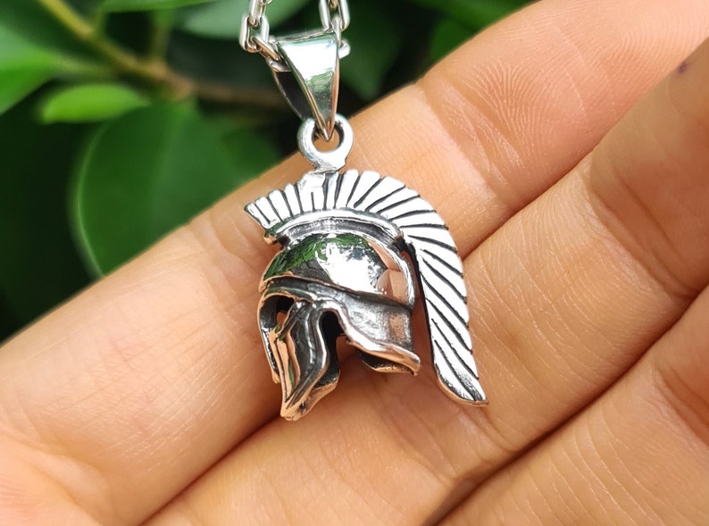 May include: Silver pendant necklace featuring a detailed Spartan helmet design. The helmet has a crested plume and a face mask.