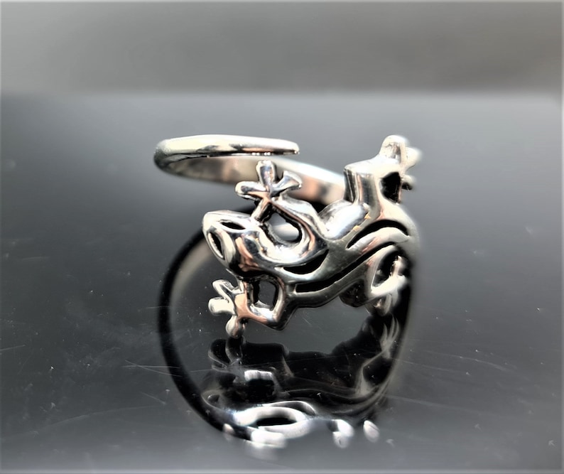 May include: A silver-colored ring featuring a dragon design. The ring has an open design with the dragon's head and body wrapping around the finger. The dragon's features are detailed, with visible eyes, mouth, and scales.
