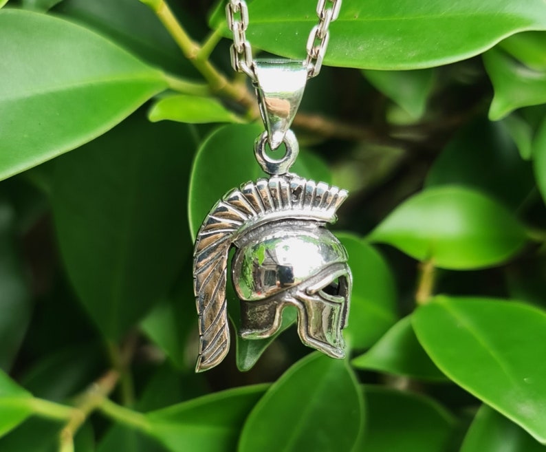 May include: Silver pendant necklace featuring a detailed, three-dimensional design of a Roman soldier's helmet. The helmet has a crested plume and a face guard.
