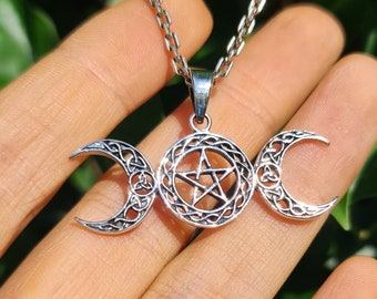May include: Silver-toned pendant necklace featuring a triple moon design with a central pentagram. The pendant has intricate Celtic knotwork and is suspended from a delicate chain. The necklace is displayed against a natural green background.