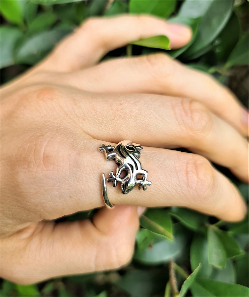May include: A silver-colored ring featuring a detailed frog design. The frog is positioned on the finger, with its legs and body wrapping around the band. The ring is set against a backdrop of green foliage.