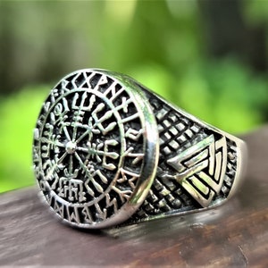 May include: A silver ring with a circular design featuring a compass with runes around the edge. The band of the ring has a Celtic knot design.