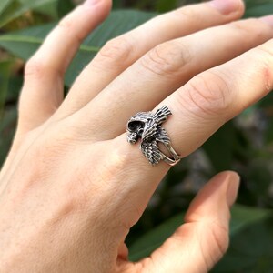May include: A silver ring featuring a detailed bird design with its wings spread out. The ring is worn on a person's finger.