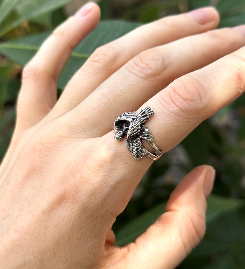 May include: A silver ring featuring a detailed bird design with its wings spread out. The ring is worn on a person's finger.