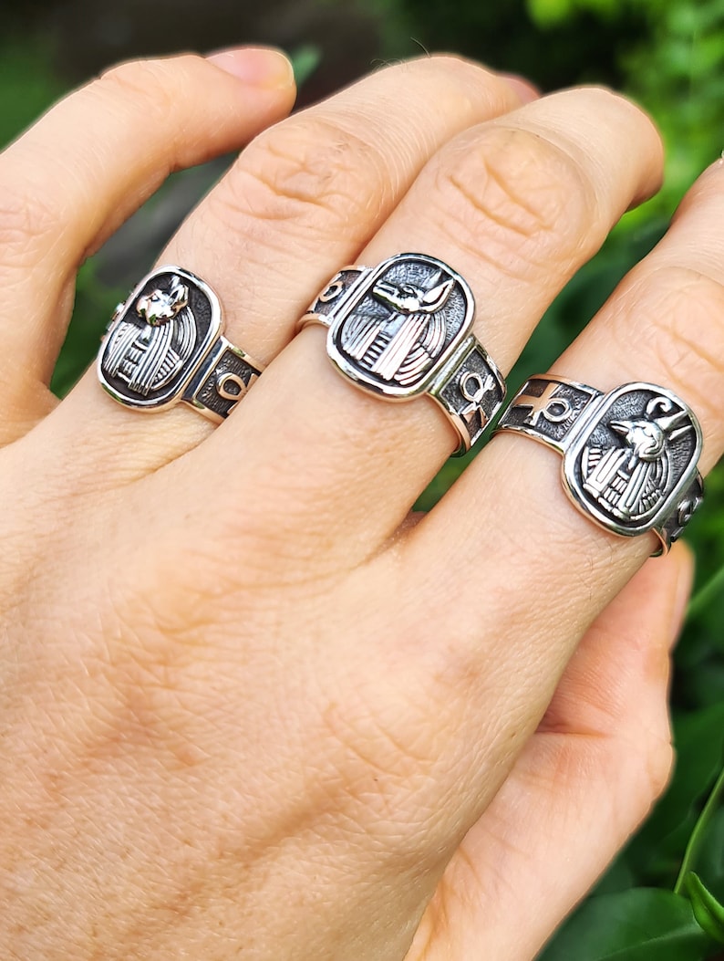 May include: Three silver rings with an Egyptian Anubis design. The rings are worn on a person's fingers.