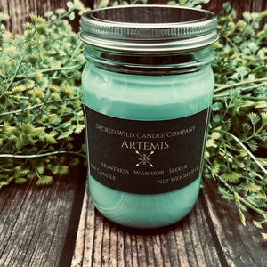 May include: A green glass jar candle with a black label that reads "Sacred Wild Candle Company Artemis Huntress Warrior Seeker Net Weight 11 oz Candle".