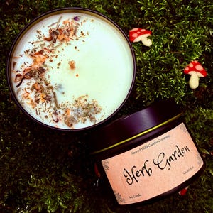 May include: A black tin candle with the text "Herb Garden" on a label. The candle has a light green wax with dried herbs and flowers. Two small mushroom decorations are in the background.