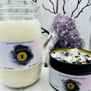 May include: Two soy candles with a black and gold label that reads "Sacred Wild Candle Company" and "Transmutation Inner Balance". The candles are in glass jars and a metal tin. The larger jar is filled with white wax and the smaller tin has a layer of white wax with blue and white crystals on top.