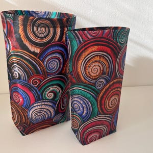 Könnte beinhalten: Zwei Stoff-Aufbewahrungstaschen mit einem farbenfrohen Spiralmuster. Die unterschiedlich großen Taschen weisen ein sich wiederholendes Design aus roten, orangefarbenen, blauen, grünen und schwarzen Spiralen auf. Die Taschen stehen aufrecht und scheinen leer zu sein.
