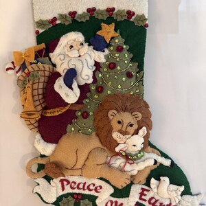 May include: A dark green felt Christmas stocking featuring an appliqued Santa Claus placing a star on a decorated tree, a lion lying with a lamb, and a white banner at the bottom with the embroidered red text Peace on Earth and a white dove.