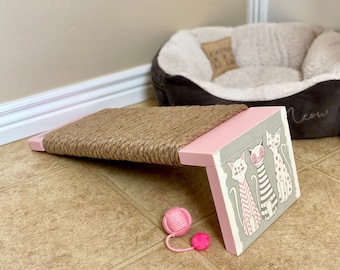 May include: Pink cat scratching post with a sisal rope surface and a side panel featuring a graphic of three cats with the word "Meow" printed on the side. A pink cat toy is on the floor.