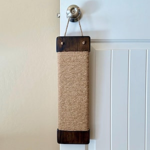 May include: A rectangular cat scratching post made of natural jute rope and dark stained wood. The post is suspended from a door knob with a jute rope. The wood frames the jute scratching surface, providing a durable and attractive design for feline scratching.