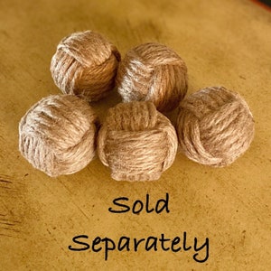 May include: Five brown, knotted, rope balls. The balls are made of natural fiber and have a rustic look. The text "Sold Separately" is written below the balls.