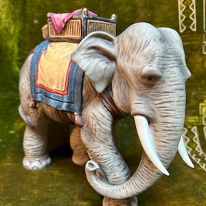Puede incluir: Figura detallada de elefante de cerámica con una silla decorativa y un pequeño carro en la espalda. El elefante es gris con colmillos de marfil y está posado sobre una superficie verde. La silla es amarilla, azul y roja.