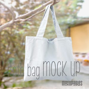 May include: A white tote bag mockup hanging from a tree branch. The bag is made of canvas and has a simple design. The text "tote bag mock up" is printed on the bag.