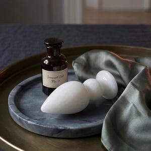 Puede incluir: Un juguete sexual blanco y liso, similar al mármol, de forma redondeada, junto a una pequeña botella marrón con una etiqueta que dice "Sensual Oil No. 3". Los artículos están sobre una bandeja de mármol gris con borde dorado y un paño de seda verde.
