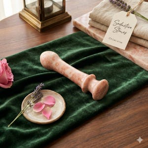 May include: A pink and white stone massage tool with the text "Seductive Stones" on a tag. The tool is 17.5 cm long and 4 cm wide. The image also includes a green velvet runner, a rose, and a small dish with lavender and rose petals.