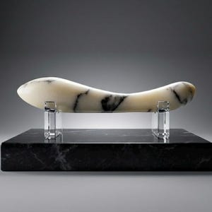 Könnte beinhalten: Eine cremefarbene, längliche Skulptur mit schwarzen Adern, die auf transparenten Acrylhaltern auf einem schwarzen Marmorsockel ausgestellt ist. Die Skulptur hat eine glatte, geschwungene Form und erzeugt eine moderne Ästhetik.