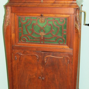 May include: A vintage wooden cabinet with a dark finish. The cabinet features ornate carvings, a green fabric panel behind a decorative wooden grille, and a hand crank. The doors have small, round knobs.